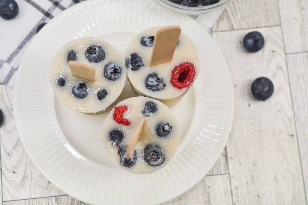 Overhead view of homemade Keto Popsicles with blueberries and raspberries on sticks, served on a white plate with a bowl of blueberries in the background.