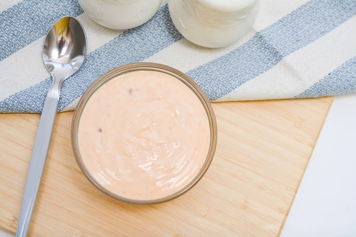 Overhead view of a bowl of Keto Bang Bang Sauce on a wooden table, with a blue cloth, a spoon, and salt and pepper shakers towards the top of the image.