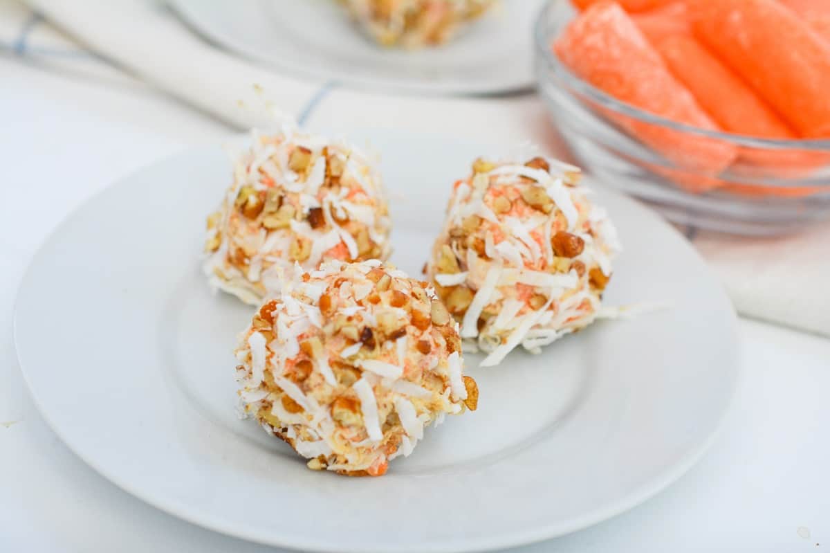 Three Keto Carrot Cake Balls served on a white plate, with a small class bowl of bright orange carrots visible in the background.