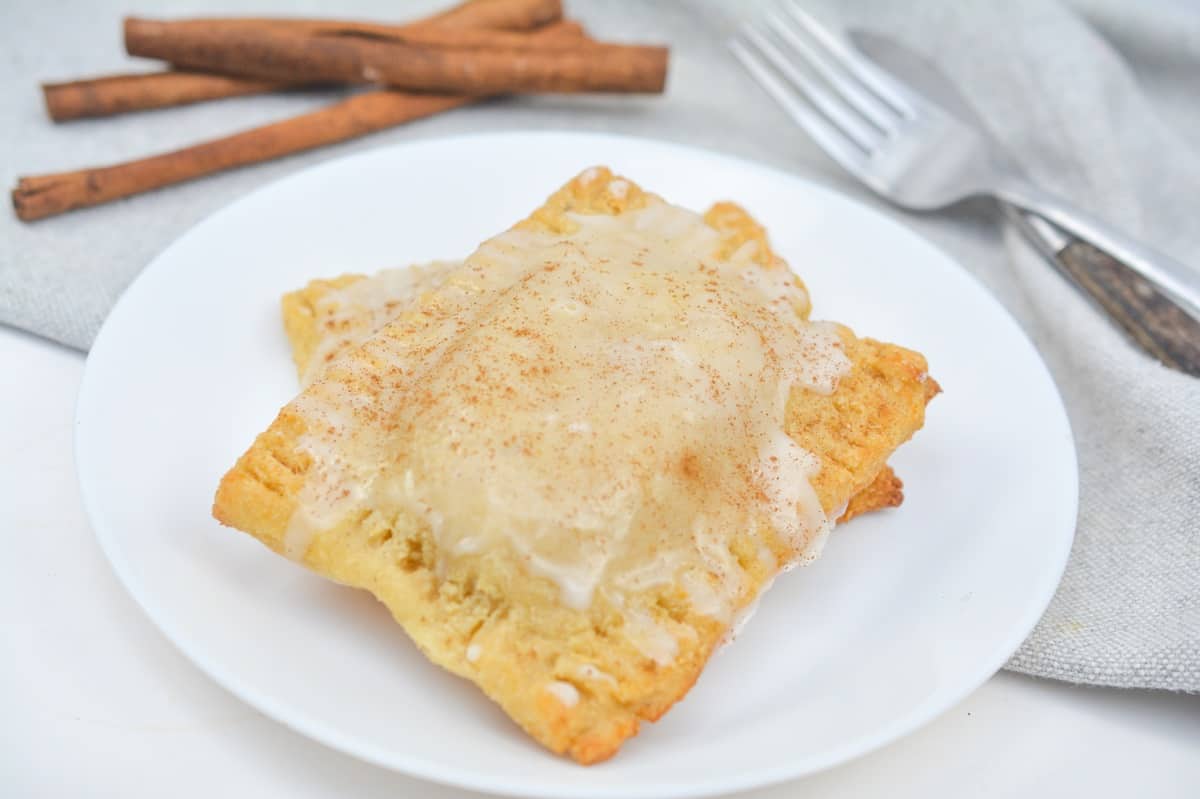 Freshly baked Keto Brown Sugar Cinnamon Pop Tarts  with icing and a sprinkle of cinnamon, served on a white plate with cinnamon sticks in the background.