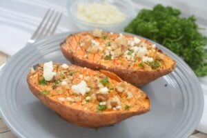 A halved Roasted Sweet Potato with Honey Garlic topped with feta cheese and chopped walnuts on a gray plate, with fresh parsley and a bowl of extra cheese in the background.
