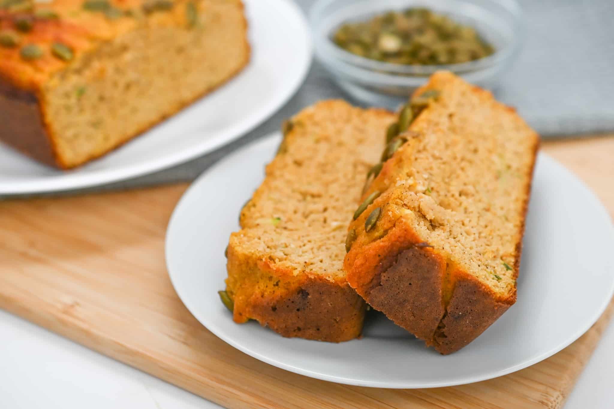 Two slices of Keto Pumpkin Zucchini Bread topped with pumpkin seeds on a white plate, placed on a wooden board with a loaf and a glass dish of pumpkin seeds in the background.