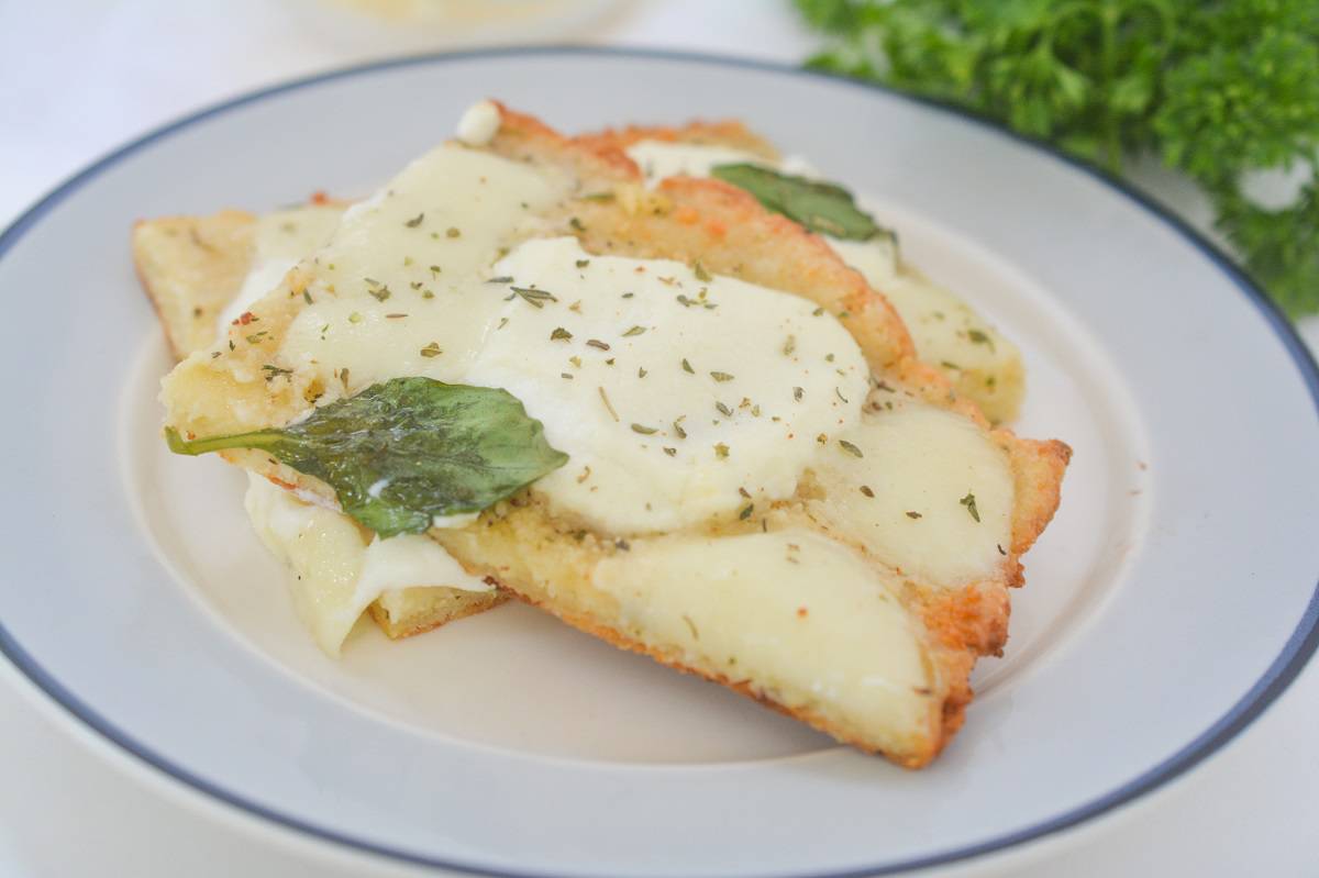Slices of Keto White Pizza garnished with fresh basil on a white plate, with grated cheese and a sprig of parsley on the side.