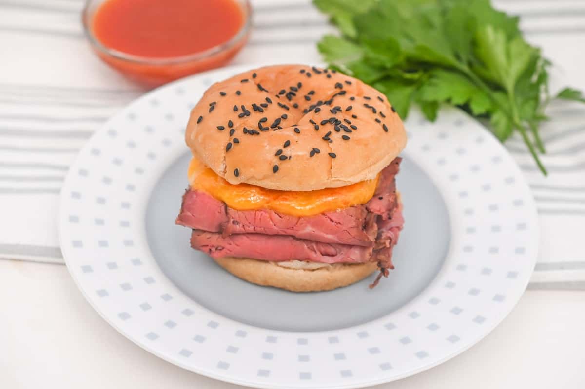 A freshly prepared Keto Copycat Arby’s Beef and Cheddar sandwich with melted cheese on a sesame seed bun, with fresh parsley as a garnish and a cup of dipping sauce.