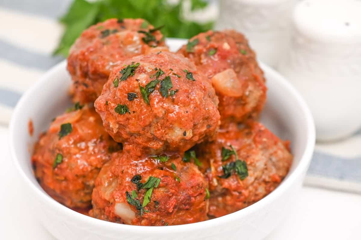 A bowl of Keto Italian Sausage Meatballs garnished with parsley, topped with a tomato-based sauce, presented on a light background with a light blue striped napkin to the top. 
