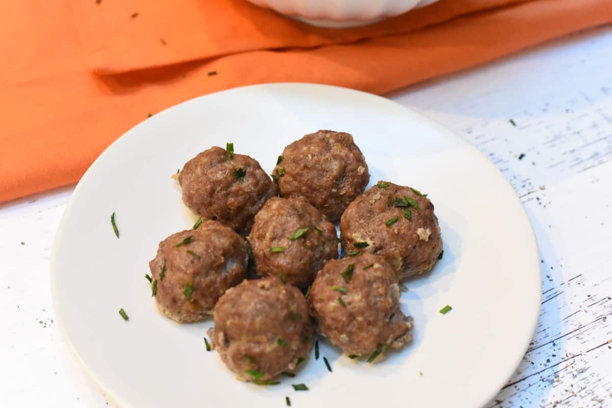 A plate of freshly-cooked Carnivore Meatballs garnished with herbs, with a bowl of more meatballs in the background atop a rustic white table and an orange napkin adding a pop of color to the setting.