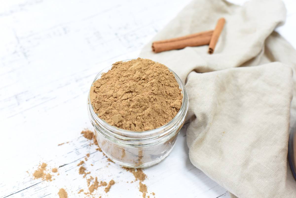 A jar filled with ingredients for making Homemade MudWtr next to two cinnamon sticks on a rustic white wooden background.