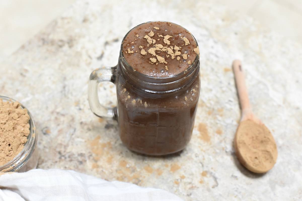 A glass jar filled with Chocolate Protein Mud Drink, sprinkled with oats on top, next to a wooden spoon with protein powder, all placed on a marble surface with a white cloth partially visible.