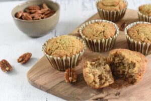 Homemade Keto Maple Pecan Muffins on a wooden board with one muffin partially unwrapped and split open, showcasing the moist crumb texture, alongside scattered pecan nuts and a bowl of additional pecans in the background, all set on a light-colored surface.