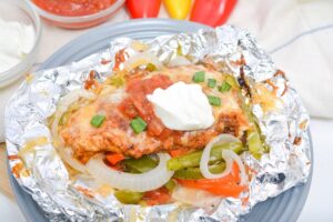 Keto Tex Mex Chicken topped with melted cheese, peppers, and onions, served on aluminum foil with a dollop of sour cream on top, displayed on a gray plate. With colorful pepper and glass bowls filled with sour cream and salsa at the top.