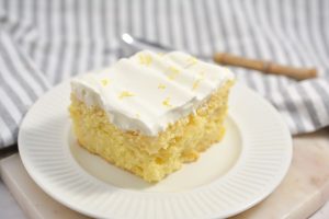 A slice of Keto Lemon Poke Cake topped with a generous layer of rich whipped cream and garnished with lemon zest on a white plate.
