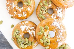 Five glazed Low Carb Gluten Free Pumpkin Spice Donuts topped with powdered sugar and pumpkin seeds are arranged on a white surface.