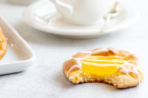 A close-up of a partially eaten lemon-filled pastry with white icing, placed on a light-colored surface. In the blurred background, there is a white teacup and saucer with a small spoon. Another pastry is partially visible on the left.