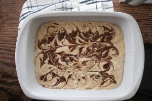 A close-up of a white baking dish filled with cream-colored batter swirled with dark brown streaks. The dish is placed on a wooden surface with a folded, white and black checkered cloth nearby.