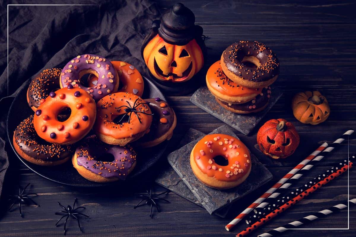 A variety of Halloween-themed doughnuts with orange and purple icing on plates, surrounded by black plastic spiders, Halloween straws, and ceramic Jack-o'-lanterns. The scene is set on a dark wooden surface with dark fabric in the background.