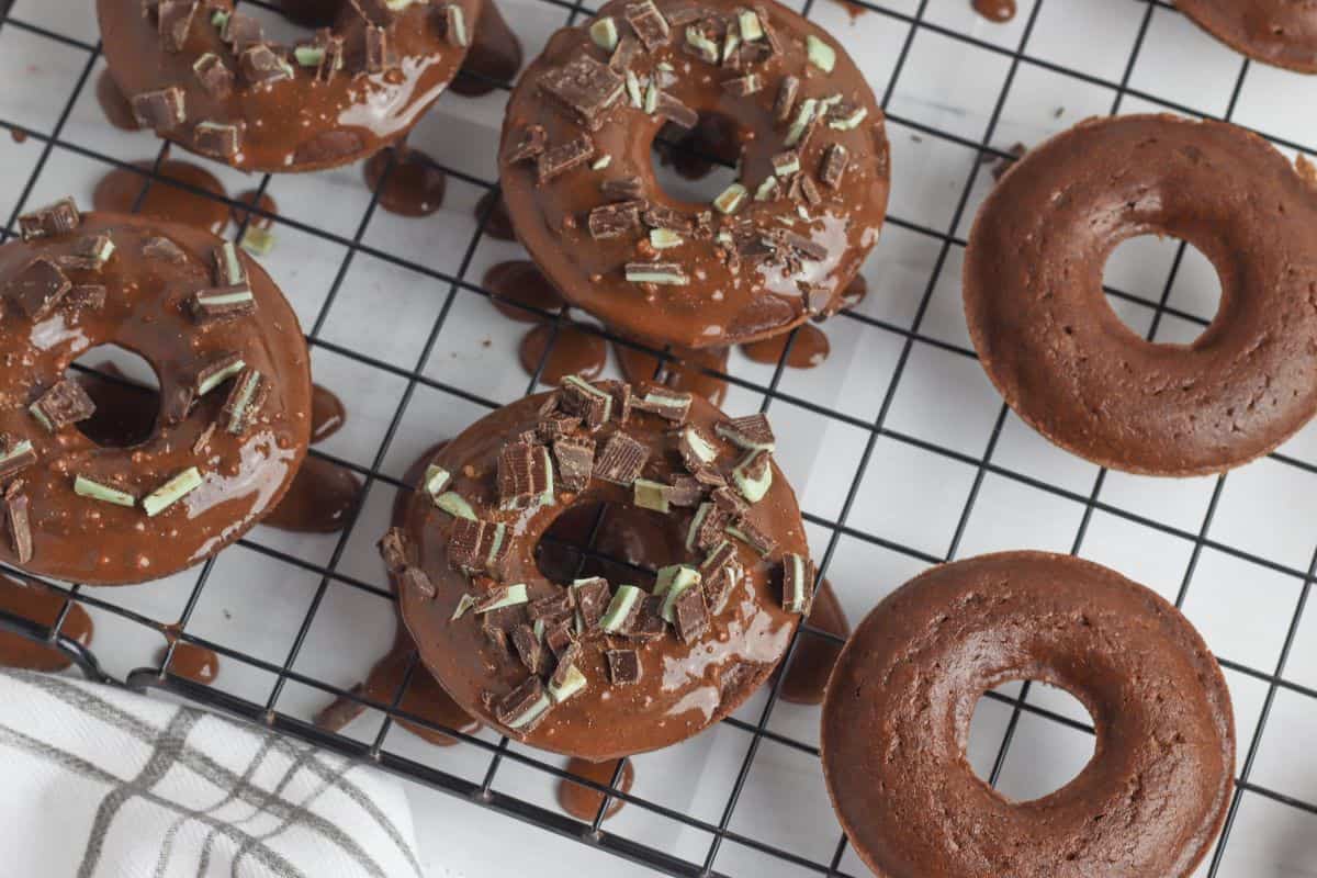 Gluten-free chocolate donuts with glossy glaze and mint chocolate bits are cooling on a black wire rack. A few donuts remain unglazed, and a white cloth with gray stripes peeks into view in the bottom left corner.