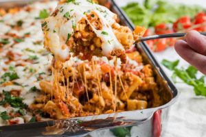 A spoon lifts a portion of cheesy baked ziti from a pan, with melted cheese stretching from the pasta. The ziti is layered with ground meat, tomato sauce, and topped with parsley. Fresh tomatoes and herbs are visible in the background.
