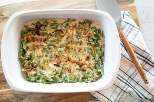 A baked green bean casserole topped with crispy onions in a white rectangular dish. The dish sits on a wooden board next to a plaid cloth napkin and a serving spoon.