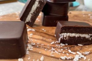 Keto Copycat Mounds Bars with a layer of white coconut filling, displayed on a wooden surface. Shredded coconut is scattered around, and more chocolate squares are in the background.