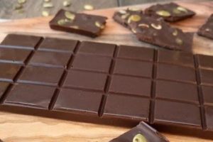 A slab of Salted Chocolate Bar with Pumpkin Seeds is displayed on a wooden board. Some pieces are broken off and scattered around the main bar, showing the textured surface and seed toppings. The background is a rustic wooden table.