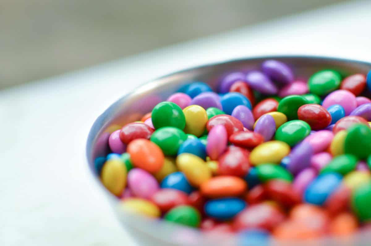 A close-up of a bowl brimming with colorful candy-coated chocolates showcases keto-friendly Halloween hacks. The candies in bright hues of red, blue, green, pink, purple, and yellow create a vibrant and cheerful display against a softly blurred background.