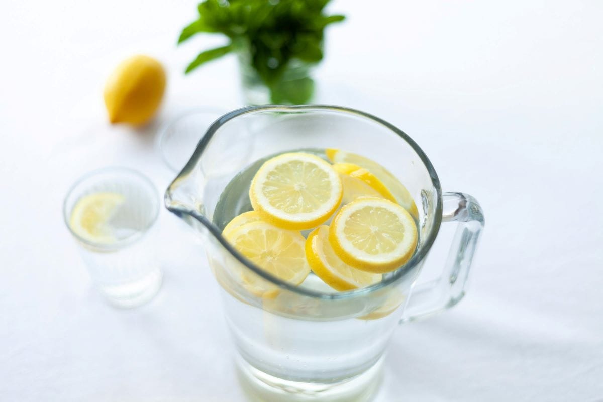 A clear glass pitcher filled with water and floating lemon slices sits on a table, offering a refreshing twist for your keto Halloween setup. In the background, a glass with a lemon slice and a vibrant yellow lemon rest near a small pot of green leaves on a white surface.