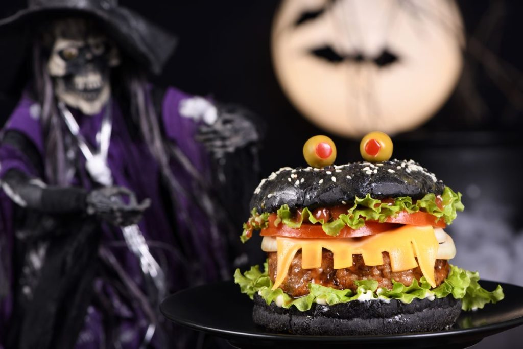 A Halloween-themed burger with a black bun, lettuce, tomato, orange cheese, and olives on top, creating a face. It sits on a black plate with a witch decoration and a blurred moon in the background. Discover Keto Halloween Hacks to make your spooky meal both festive and keto-friendly!