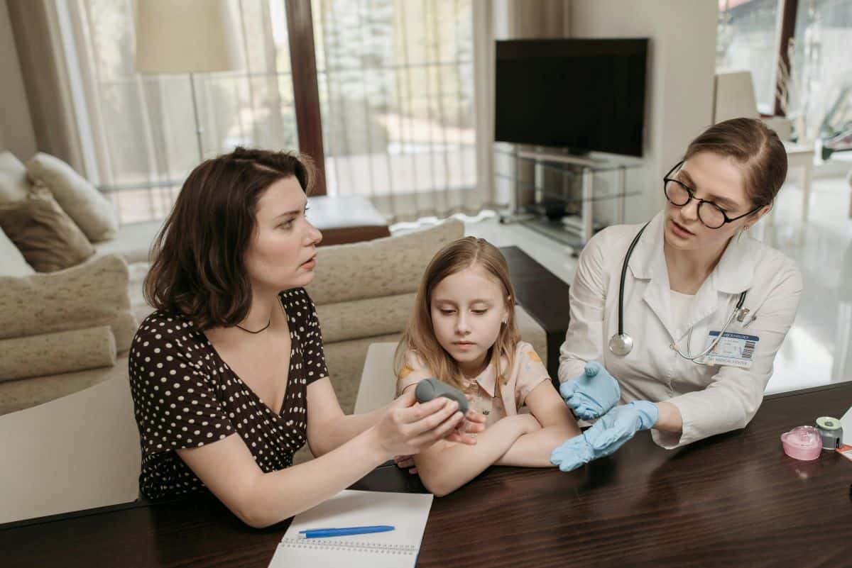 A woman and a child sit at a table with a female doctor who is wearing a white coat and gloves. The woman holds a small black device, while the doctor appears to be explaining something, possibly about keto Halloween hacks. A notepad and pen are on the table.