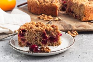 A slice of cranberry walnut crumb cake on a plate, showcasing visible cranberries and walnuts. Perfect among sweet loaves and breakfast cakes, it sits against a wooden board with the remaining cake. A fork and a white napkin accompany it on the gray surface.