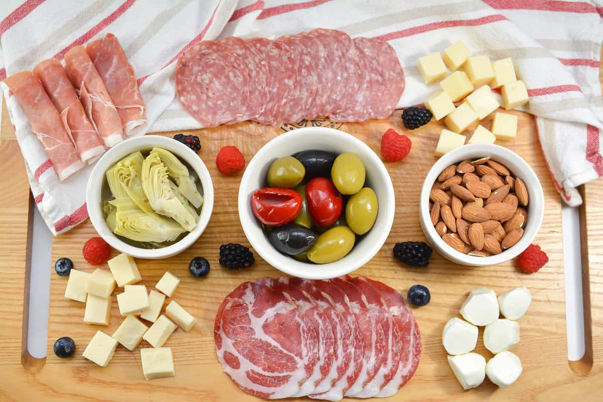 A charcuterie board with cured meats, olives, artichoke hearts, almonds, cheese cubes, mozzarella balls, and assorted berries on a wooden surface with a striped cloth.