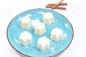 A blue plate with six white, flower-shaped desserts arranged neatly. Three cinnamon sticks are placed in the background on a white textured surface.