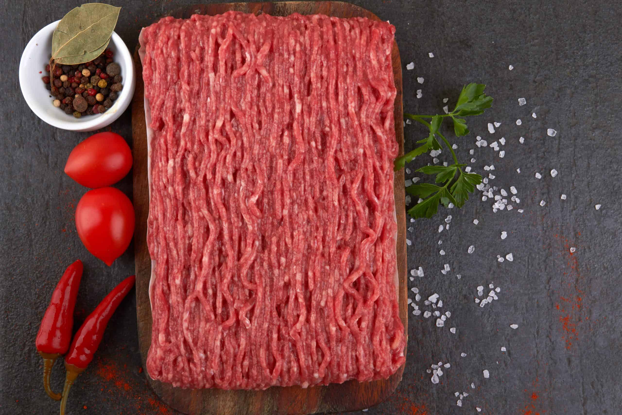 A slab of raw ground beef on a wooden board is surrounded by fresh tomatoes, red chili peppers, and coarse salt. A small bowl of mixed peppercorns and a bay leaf is also nearby, with sprigs of fresh parsley on a dark textured surface.