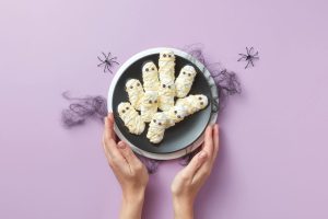 Hands hold a black plate with Halloween-themed mummy cookies wrapped in white icing and candy eyes. The plate rests on a white dish against a purple background, decorated with black cobwebs and small spider cutouts.