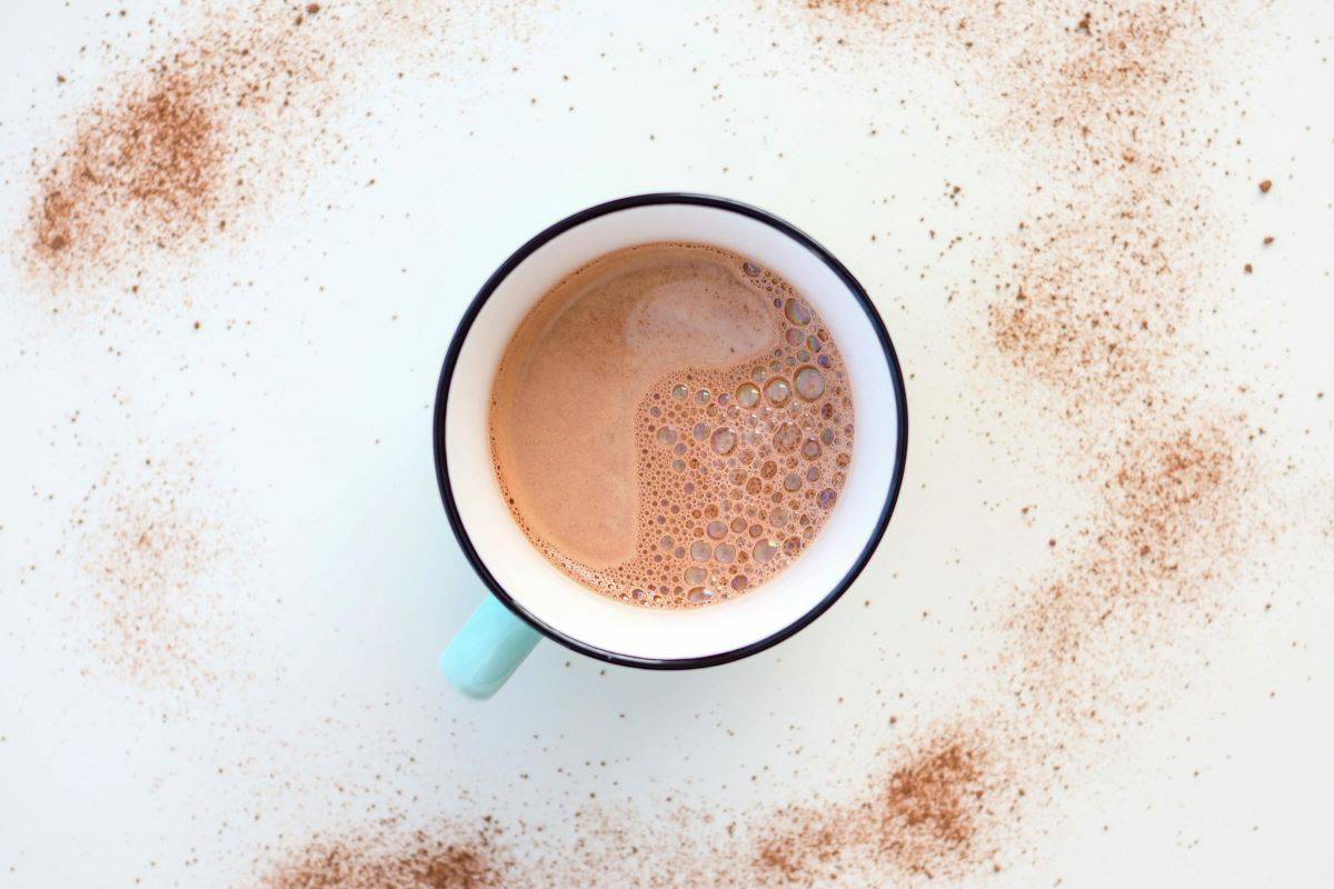 A cup of hot chocolate with frothy bubbles on the surface, placed on a white background. Cocoa powder is artistically sprinkled around the cup, creating a textured effect. The cup has a light blue handle.