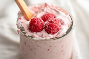 A glass jar of creamy, no-sugar raspberry yogurt topped with three fresh raspberries. A wooden spoon is sticking out of the yogurt, and the jar is placed on a pristine white surface.