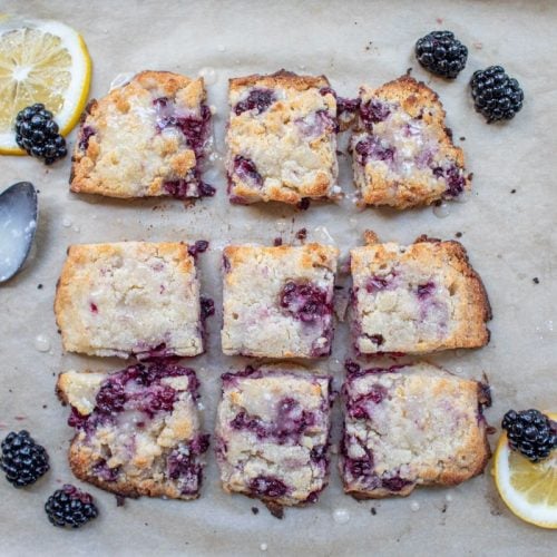 A tray lined with parchment paper holds nine square pieces of blackberry crumb bars, surrounded by fresh blackberries and lemon slices. A spoon with a bit of icing and a small container of icing are on the left side of the tray.