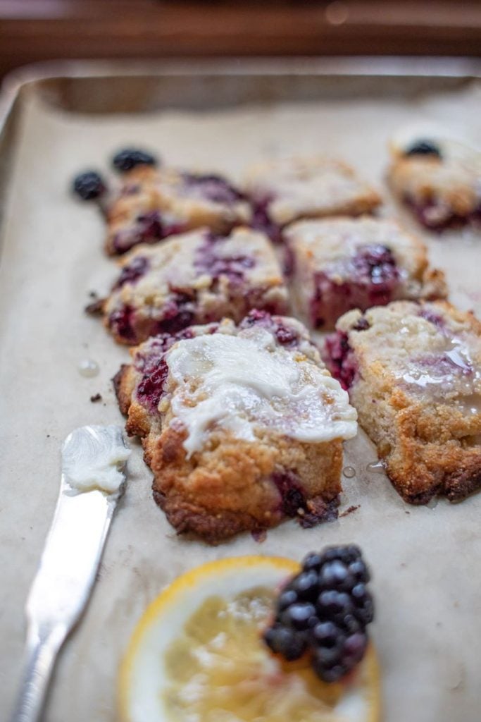 Close-up of blackberry scones on a baking sheet. The scones are topped with a light glaze and some blackberries are mixed into the dough. A knife with some glaze on it and a slice of lemon garnished with blackberries are placed in the foreground.