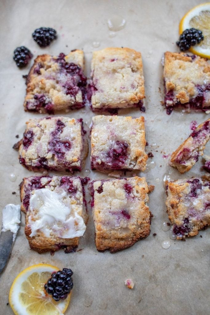 Nine square pieces of blackberry lemon cake, arranged on parchment paper. The cake pieces are topped with icing drizzle and surrounded by fresh blackberries and lemon slices. A butter knife with a bit of white spread lies next to one of the pieces.
