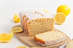 A lemon loaf cake with icing rests on a wooden board, embodying the essence of sweet loaves. A slice is cut and placed in front, while whole and sliced lemons add brightness to the scene. The background features a light, neutral color that complements this delightful breakfast cake.