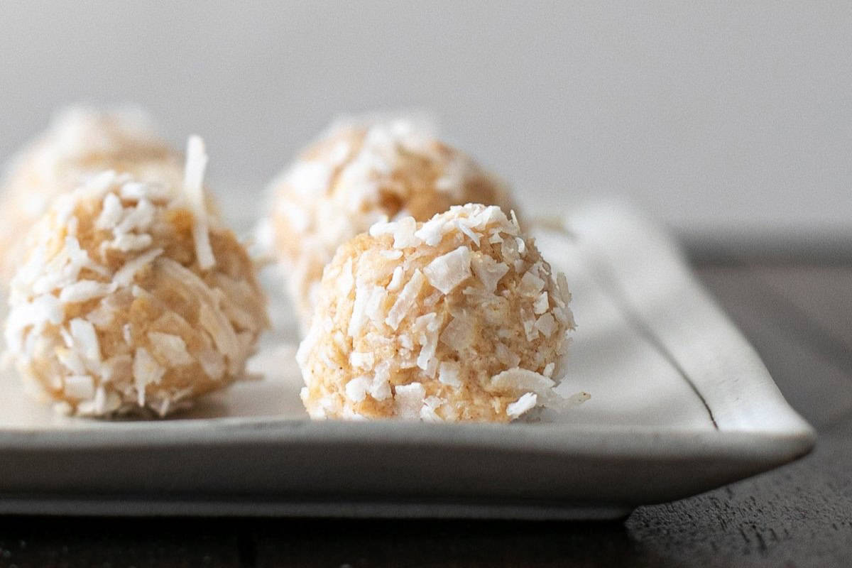 Close-up of a rectangular white plate with three round, light brown coconut truffles coated in shredded coconut. The truffles are in focus, while the plate's edge and background are softly blurred.