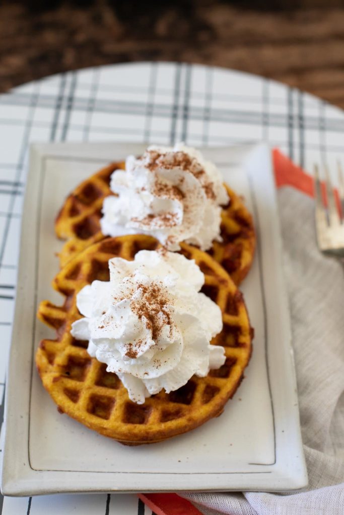 Two pumpkin waffles topped with whipped cream and a sprinkle of cinnamon are served on a rectangular white plate. The setting includes a patterned tablecloth, a red napkin, and a fork.