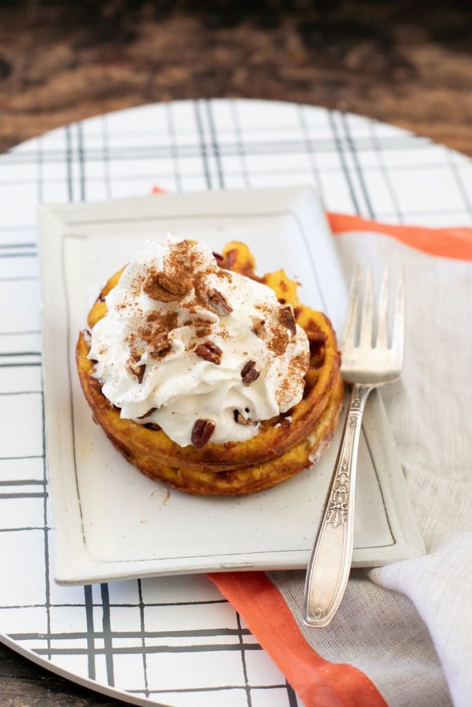 A waffle topped with whipped cream, sprinkled with cinnamon and pecans, is served on a rectangular white plate. A vintage fork rests beside the waffle, on a patterned tablecloth with orange accents.