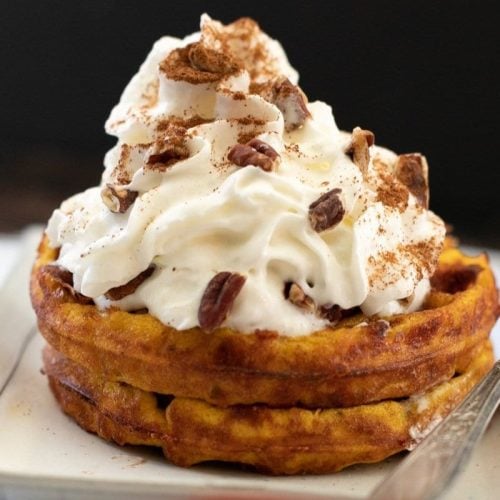 A stack of pumpkin-flavored waffles on a rectangular plate, topped with a generous swirl of whipped cream, sprinkled with cinnamon, and garnished with pecans. A fork is placed beside the waffles.