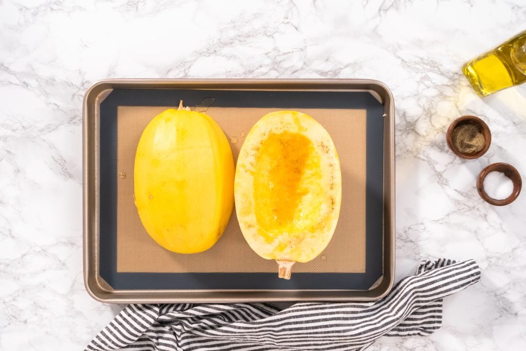 A baking tray on a marble surface holds two spaghetti squash halves, one seeded, one whole. Nearby are a bottle of olive oil and two small wooden bowls. A black and white striped cloth lies at the bottom of the tray.