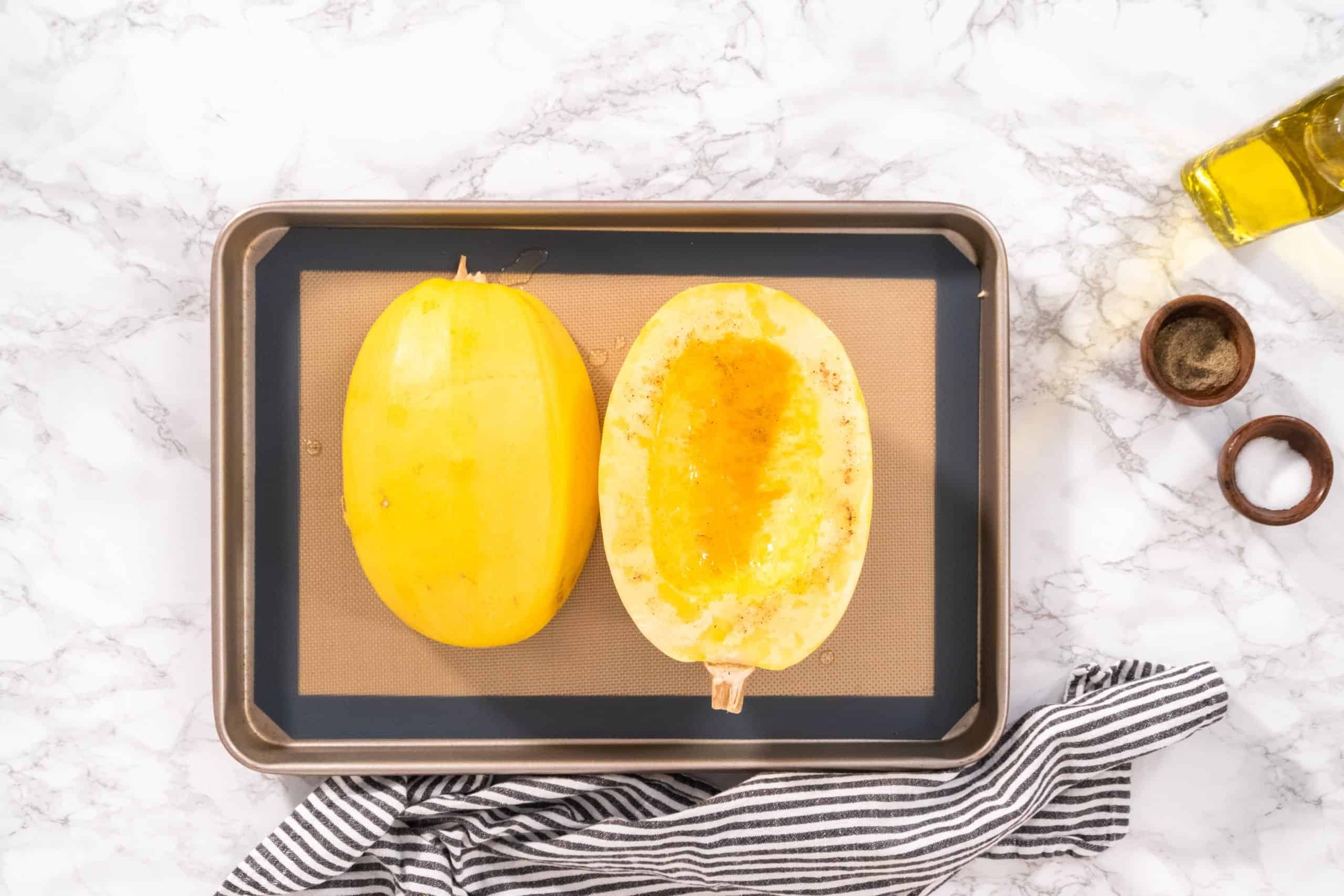 A baking tray on a marble surface holds two spaghetti squash halves, one seeded, one whole. Nearby are a bottle of olive oil and two small wooden bowls. A black and white striped cloth lies at the bottom of the tray.