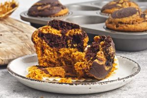 A gluten-free marble muffin, featuring swirls of chocolate and pumpkin, is split open on a white plate. In the background, more muffins rest in a baking tray, highlighting the mix of rich brown and vibrant orange colors in the batter.