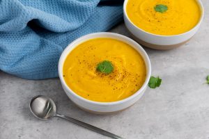 Two bowls of creamy carrot soup garnished with a sprig of cilantro and black pepper sit on a gray surface. A silver spoon and a blue textured cloth are nearby.