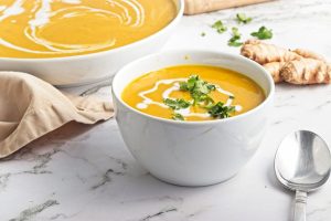 A white bowl filled with creamy orange soup garnished with cilantro and a drizzle of cream. Fresh ginger root and a matching bowl of soup are in the background, all on a marble countertop. A silver spoon and a beige napkin are nearby.