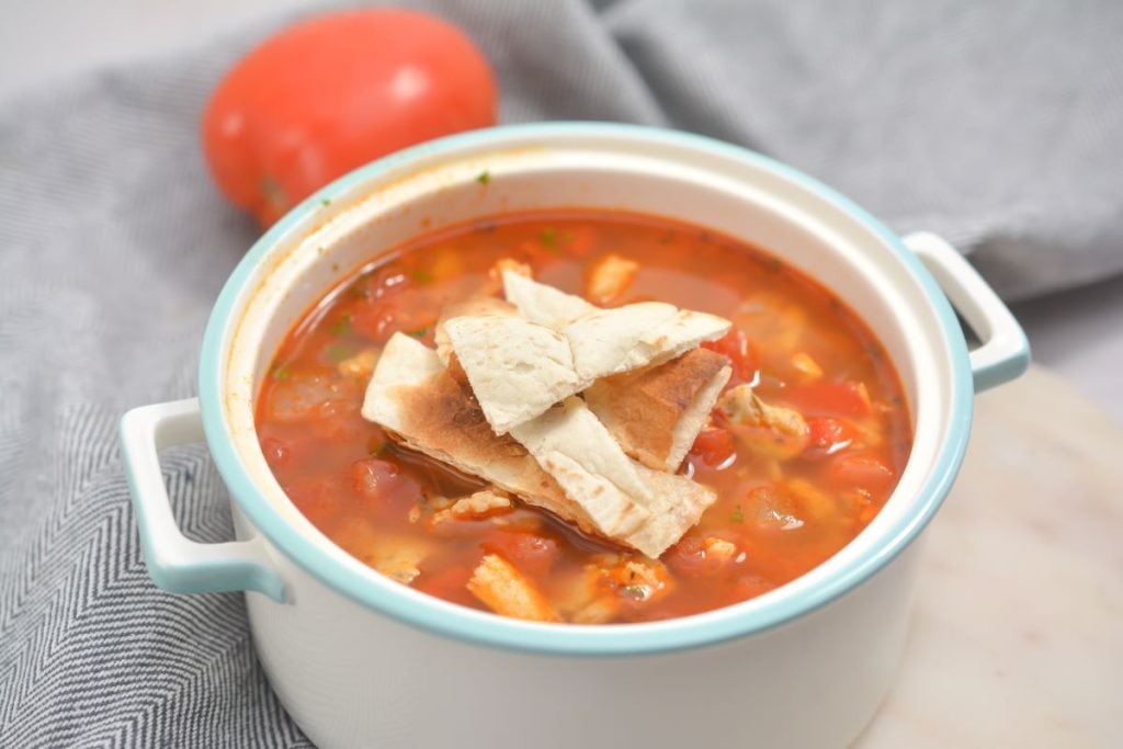A bowl of hearty tomato soup topped with crispy bread pieces, placed on a white surface. A whole tomato is in the background, partially visible on a gray cloth.