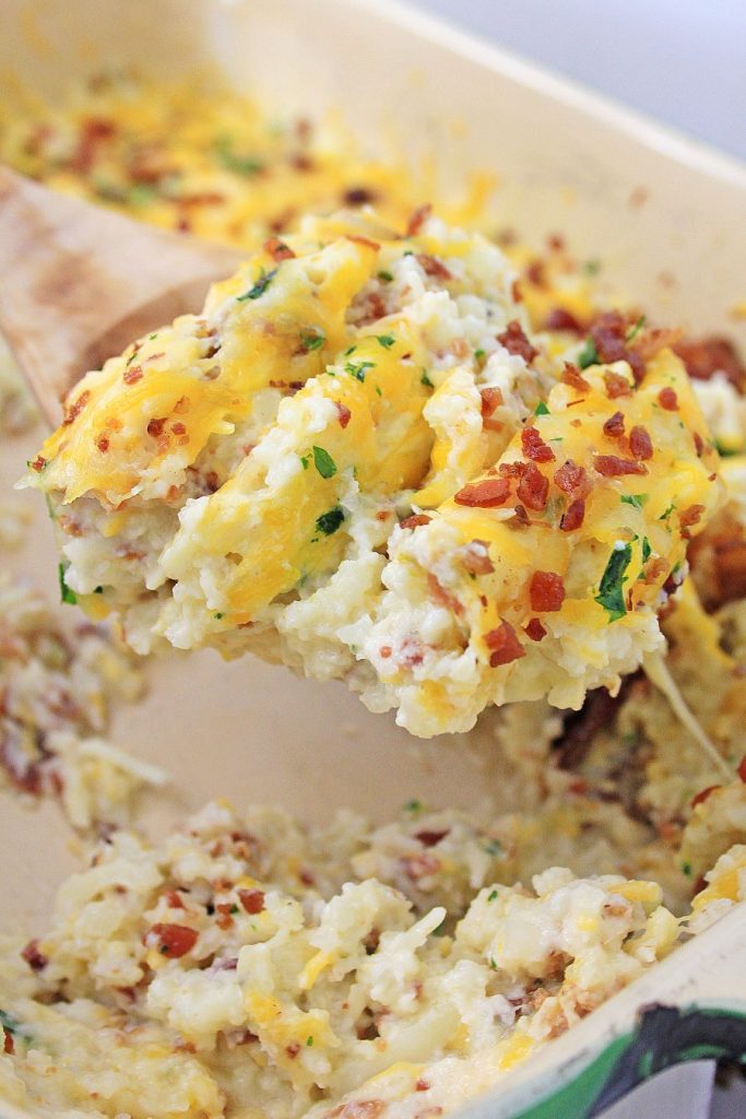 A serving of creamy mashed cauliflower loaded with melted cheese, crispy bacon bits, and chopped herbs is being lifted from a baking dish with a wooden spoon. The dish is garnished with fresh herbs and crumbled bacon.